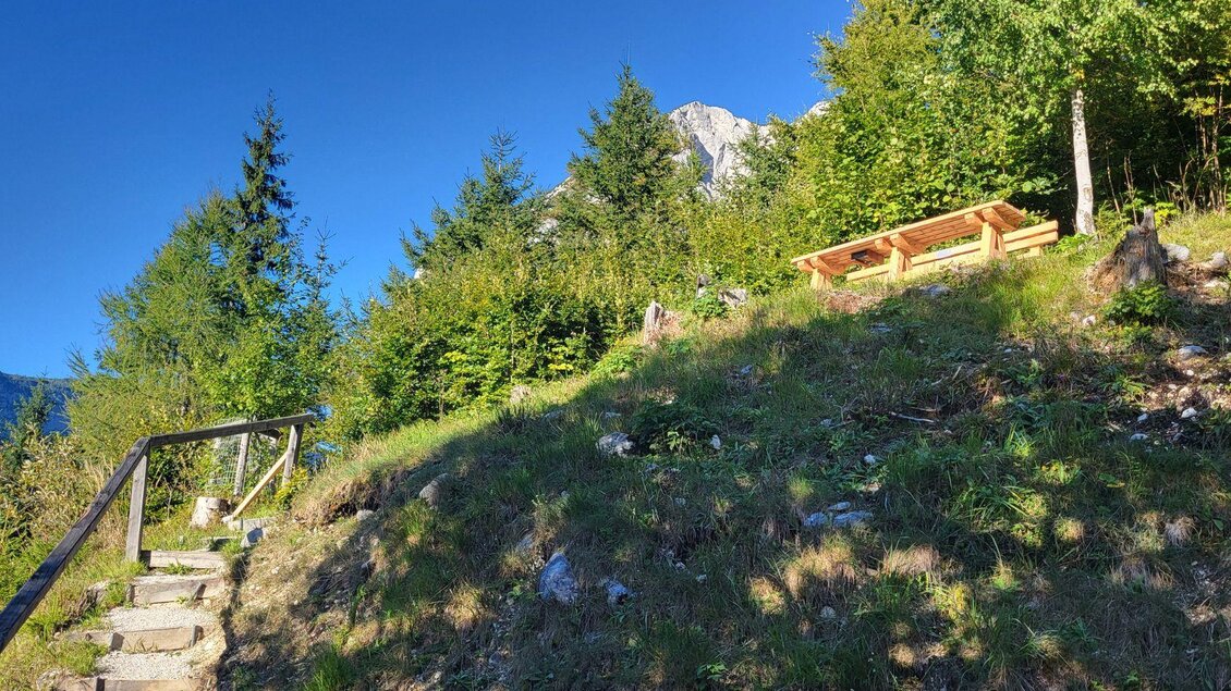 Ein Weg führt durch eine grüne Wiese zu einer Holzbank im Freien. Im Hintergrund sind Bäume und Berge zu sehen. | © TVB Ausseerland Salzkammergut_Petra Kirchschlager