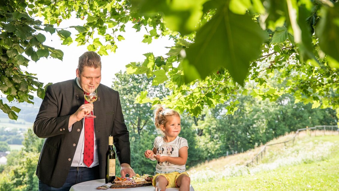 Ein Mann in einem Anzug genießt einen Drink, während ein kleines Mädchen an einem Tisch mit Snacks sitzt. Im Hintergrund ist eine grüne Landschaft unter einem Baum zu sehen. | © TVB Bad Waltersdorf