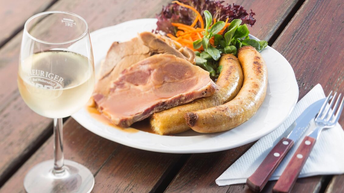 Ein Teller mit Bratwurst, Fleisch und frischem Salat. Dazu steht ein Glas Weißwein auf einem Holztisch. | © Bernhard Bergmann