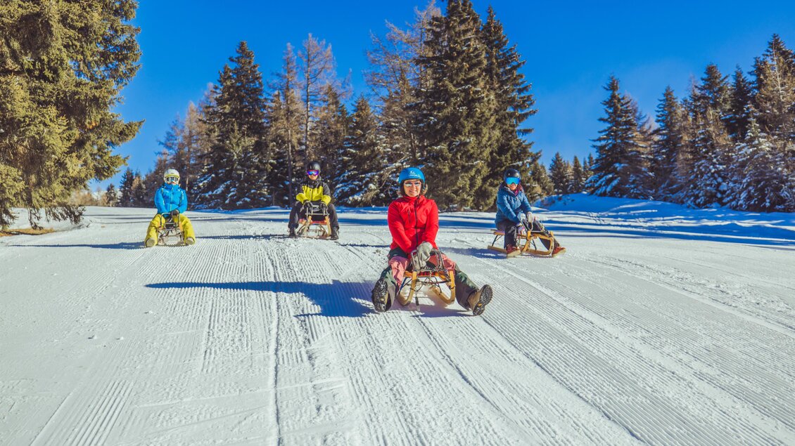 Eine Gruppe von Kindern fährt mit Schlitten auf einer verschneiten Piste. Im Hintergrund sind verschneite Bäume und ein klarer blauer Himmel zu sehen. | © Tourismusverband Murau