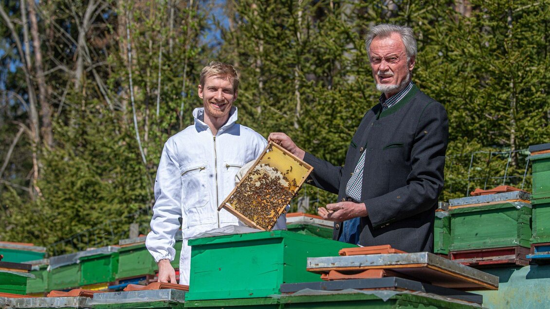 Zwei Männer stehen vor Bienenstöcken im Freien. Einer von ihnen hält eine Wabe mit Bienen in der Hand. | © Jürgen Fuchs