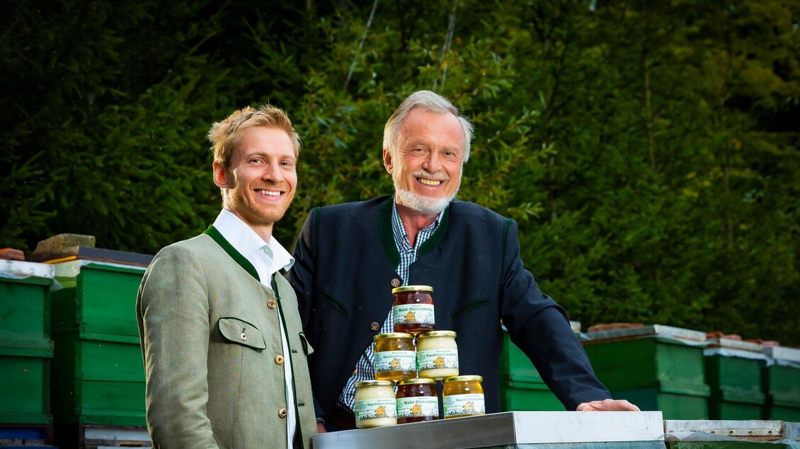 Zwei Männer stehen vor Bienenstöcken und lächeln in die Kamera. Auf einem Tisch vor ihnen sind Gläser mit Honig gestapelt. | © Gruber