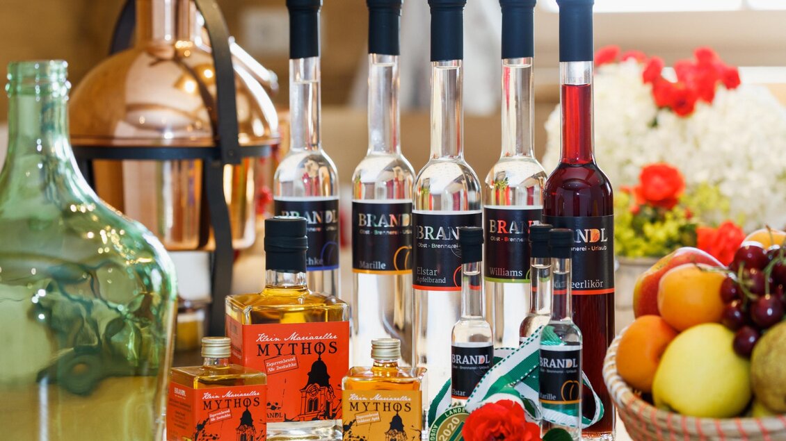 Eine Auswahl an verschiedenen Spirituosenflaschen auf einem Tisch. Im Hintergrund sind einige Früchte und Blumen zu sehen. | © Familie Brandl