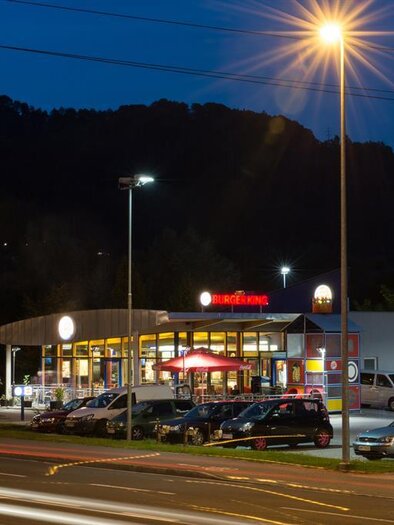 A Burger King restaurant at night, illuminated by street lamps. Numerous cars are parked in front of the establishment. | © Fotostudio Helmut Reisinger
