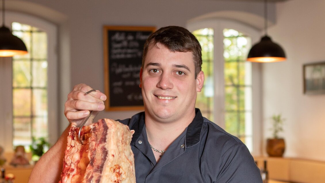 Ein Mann hält ein großes Stück Fleisch in der Hand und lächelt stolz. Im Hintergrund ist ein gemütliches Restaurant zu sehen. | © GlaMUR-Genuss am Fluss