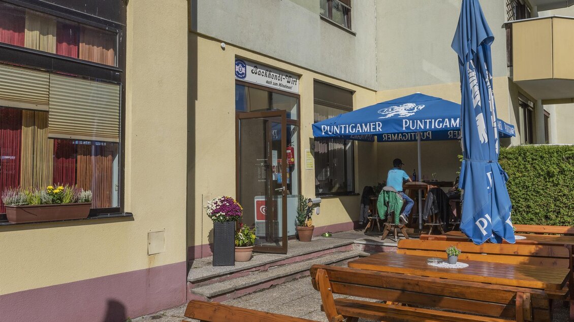 Ein einladendes Restaurant mit einer Außenbestuhlung aus Holz und einem blauen Sonnenschirm. Der Eingang ist freundlich gestaltet und es gibt Blumen im Blumenkasten. | © Familie Mutlu