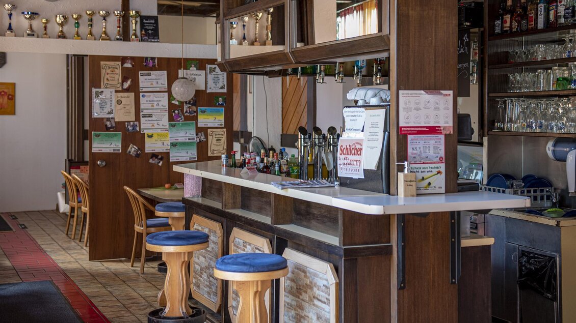 Eine gemütliche Bar mit einer langen Theke und blauen Barhockern. Im Hintergrund sind Regale mit Getränken und Auszeichnungen zu sehen. | © Familie Mutlu