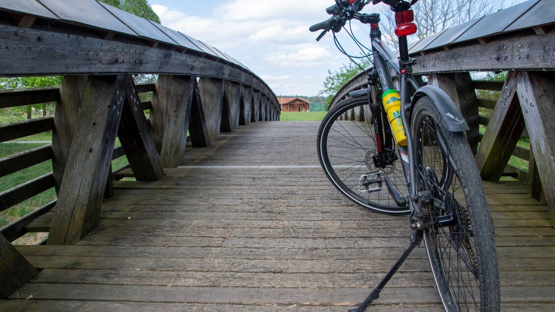 Ein Fahrrad steht auf einer Holzbrücke mit einem schönen, grünen Hintergrund. Der Himmel ist klar und es gibt wenig Wolken. | © Kurkommission Bad Blumau
