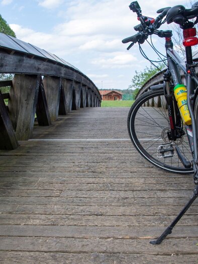 Ein Fahrrad steht auf einer Holzbrücke mit einem schönen, grünen Hintergrund. Der Himmel ist klar und es gibt wenig Wolken. | © Kurkommission Bad Blumau