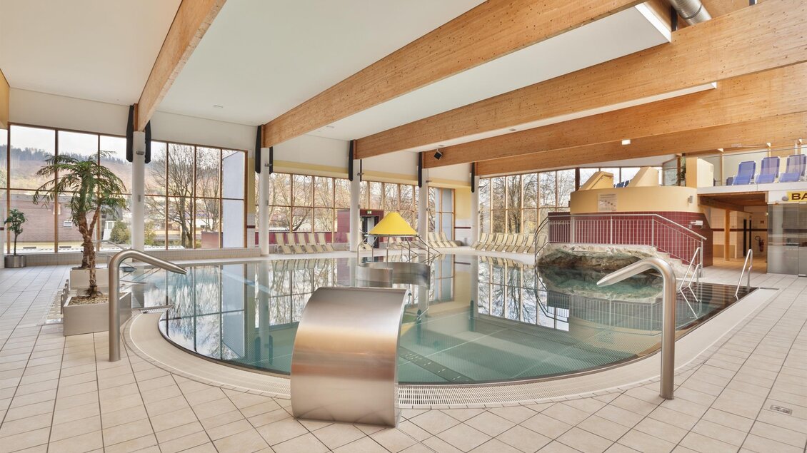 Ein modernes Hallenbad mit einem großen Becken und großen Fenstern. Die Holzdecken verleihen dem Raum eine warme Atmosphäre.