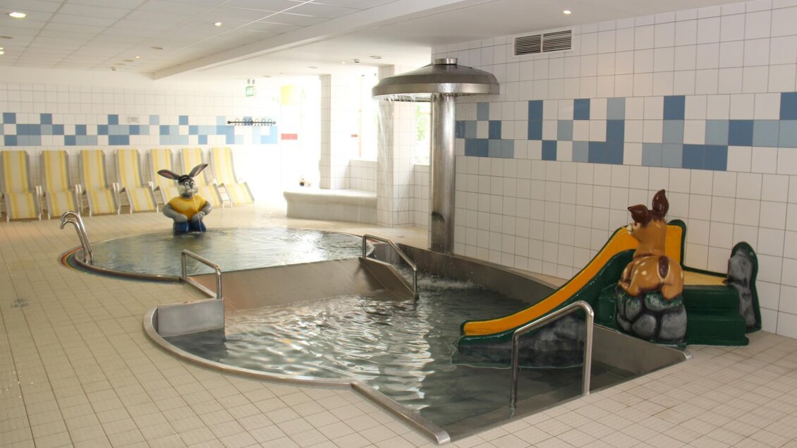 Ein Innenpool mit einer rutschigen Rutsche und einem Wasserspielzeug. Umgeben von Liegen und einer hellen, freundlichen Umgebung.