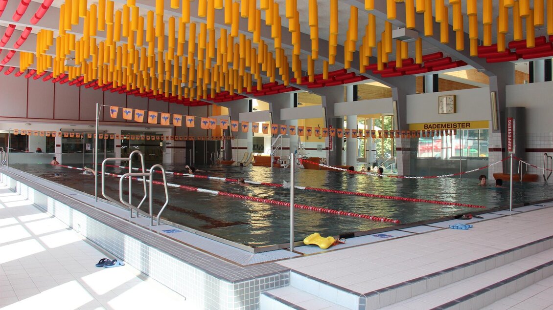 Ein helles Schwimmbad mit Wasser und Schwimmbahnen. Bunte Akzente an der Decke geben dem Raum eine fröhliche Atmosphäre.