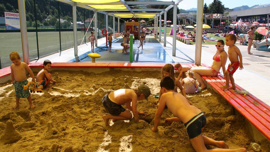 Eine sandige Spielzone, in der Kinder fröhlich im Sand spielen. Im Hintergrund sind Sonnenschirme und Kinder in Badesachen zu sehen.
