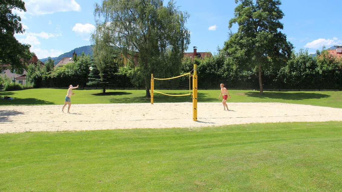 Ein Sandvolleyballplatz in einem grünen Park. Zwei Personen spielen Volleyball unter einem klaren blauen Himmel.