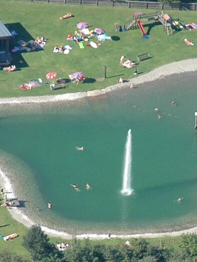 A beautiful pond with a water feature and many sun lounges. Around the pond, numerous people and colorful sun umbrellas can be seen. | © Gemeinde Donnersbach
