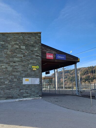 A modern train station with a stone facade. The sky is clear and the surroundings show trees and tracks. | © Heidi Grundner