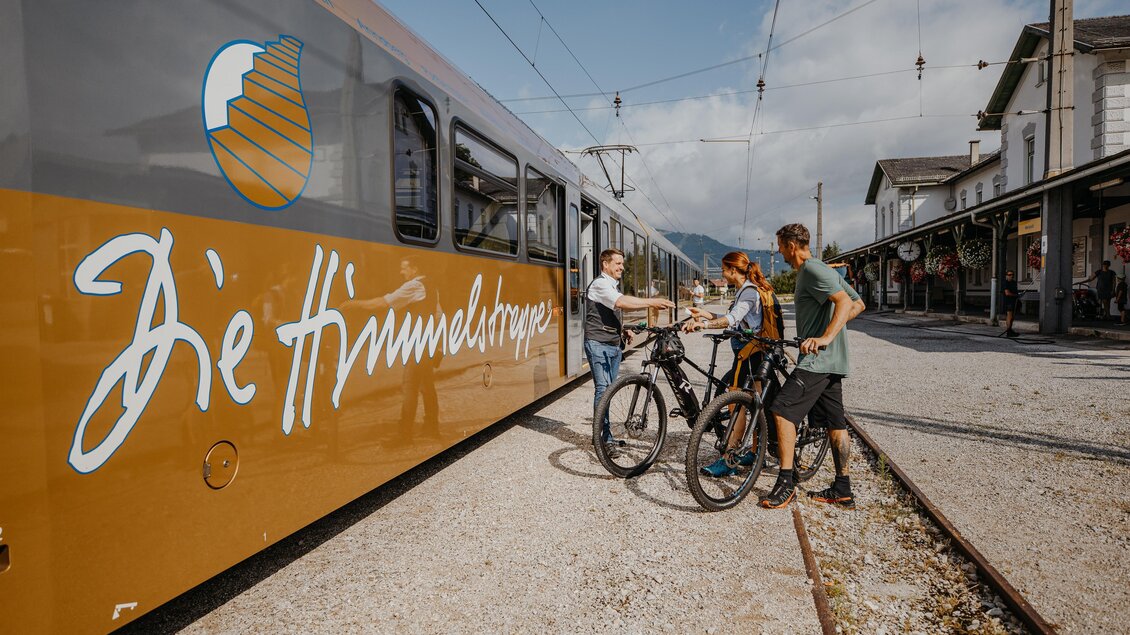 Eine Gruppe von Radfahrern steht neben einem gelben Zug. Im Hintergrund sind Gleise und ein Bahnhof zu sehen. | © TV Hochsteiermark, nicoleseiser.at
