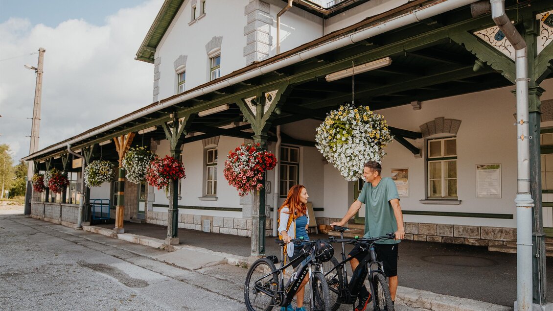 Ein altes Bahnhofsgebäude mit blühenden Pflanzen und zwei Fahrrädern. Zwei Personen stehen entspannt davor und genießen die Atmosphäre. | © TV Hochsteiermark, nicoleseiser.at