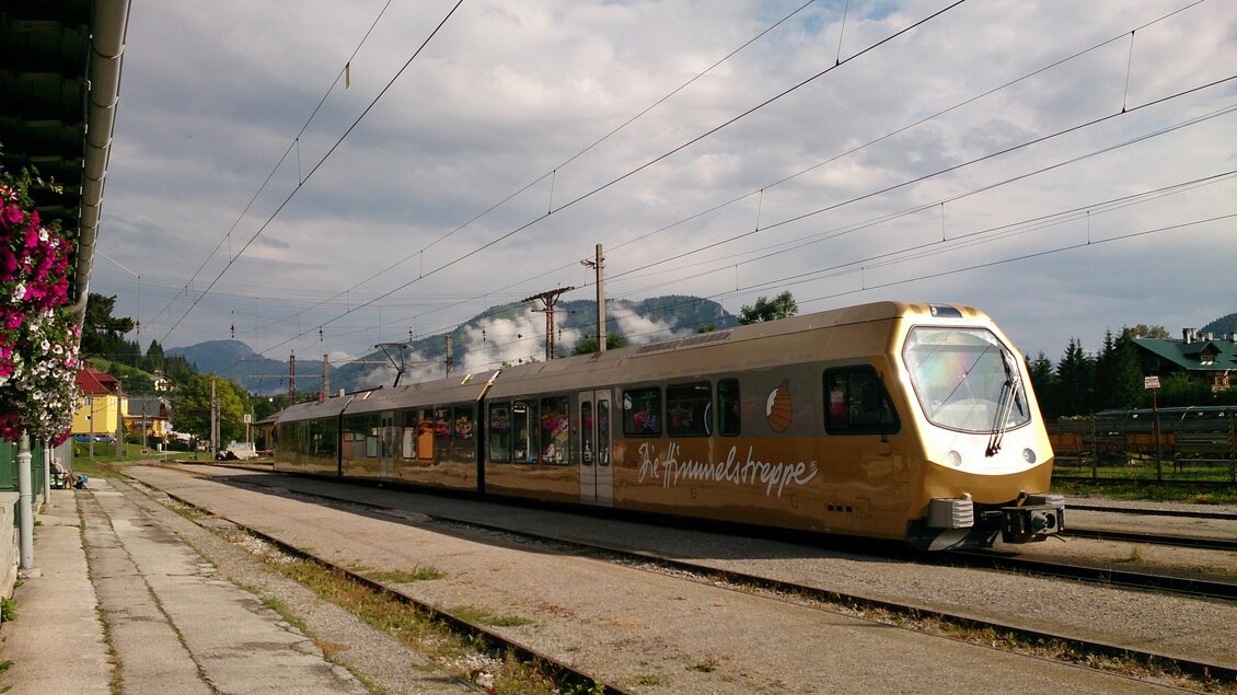 Ein modern aussehender Zug steht auf einem Bahnhof, umgeben von einer ländlichen Landschaft. Im Hintergrund sind Berge und ein bewölkter Himmel sichtbar. | © TV Hochsteiermark