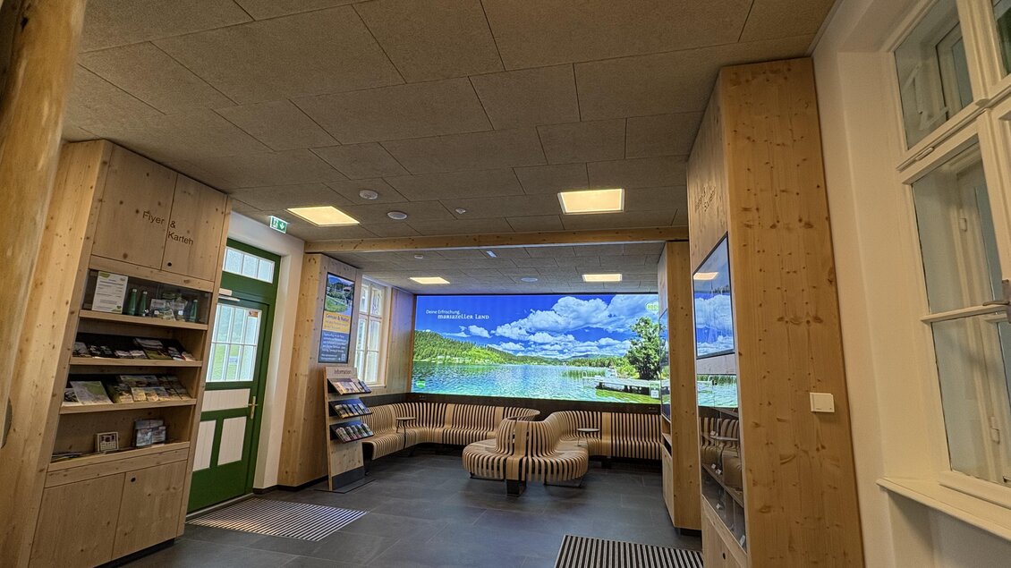 Ein moderner Empfangsbereich mit Holzvertäfelung und einem großen Bildschirm, der eine malerische Landschaft zeigt. Gemütliche Sitzgelegenheiten laden zum Verweilen ein. | © TV Hochsteiermark
