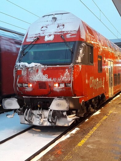 Ein roter Zug steht in einem verschneiten Bahnhof. Der Schnee bedeckt die Gleise und die Umgebung ist winterlich. | © Symbolfoto