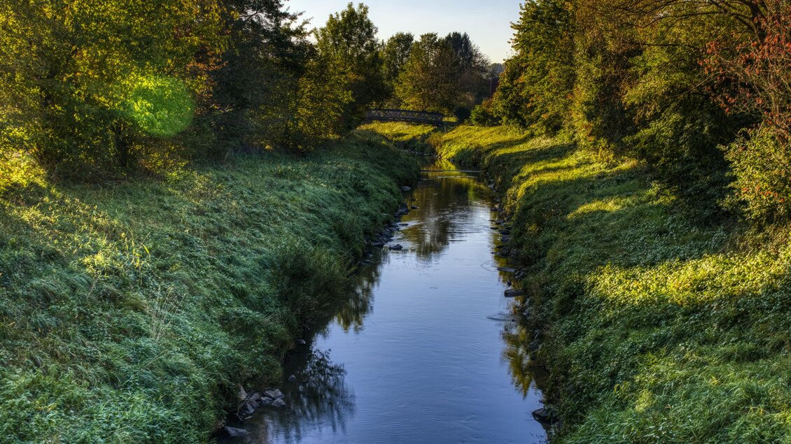 Ein ruhiger Wasserlauf umgeben von üppigem Grün und Bäumen. Der Himmel ist klar und die Atmosphäre wirkt friedlich. | © Kurkommission Bad Blumau