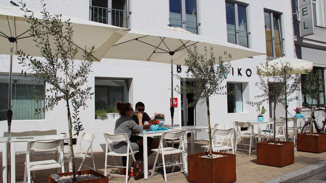 Ein modernes Restaurant mit einer gemütlichen Außenterrasse. Es gibt weiße Tische, Sonnenschirme und grüne Pflanzen. | © Bakaliko