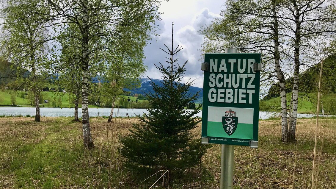Ein Schild weist auf ein Naturschutzgebiet hin. Im Hintergrund sind Bäume und ein Teich zu sehen. | © Sabine Schulz