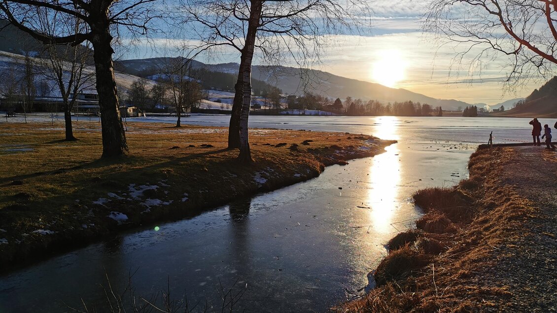 Ein ruhiger Fluss bei Sonnenuntergang, umgeben von Bäumen und Bergen. Die Oberfläche des Wassers spiegelt das warme Licht der Sonne wider. | © Sabine Schulz