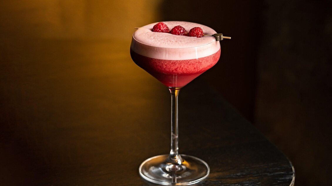 Ein eleganter Cocktail in einem hohen Glas, garniert mit frischen Himbeeren. Die Farbgebung ist lebhaft rot mit einer schaumigen, helleren Schicht oben. | © Bar Amouro - Lucija Novak