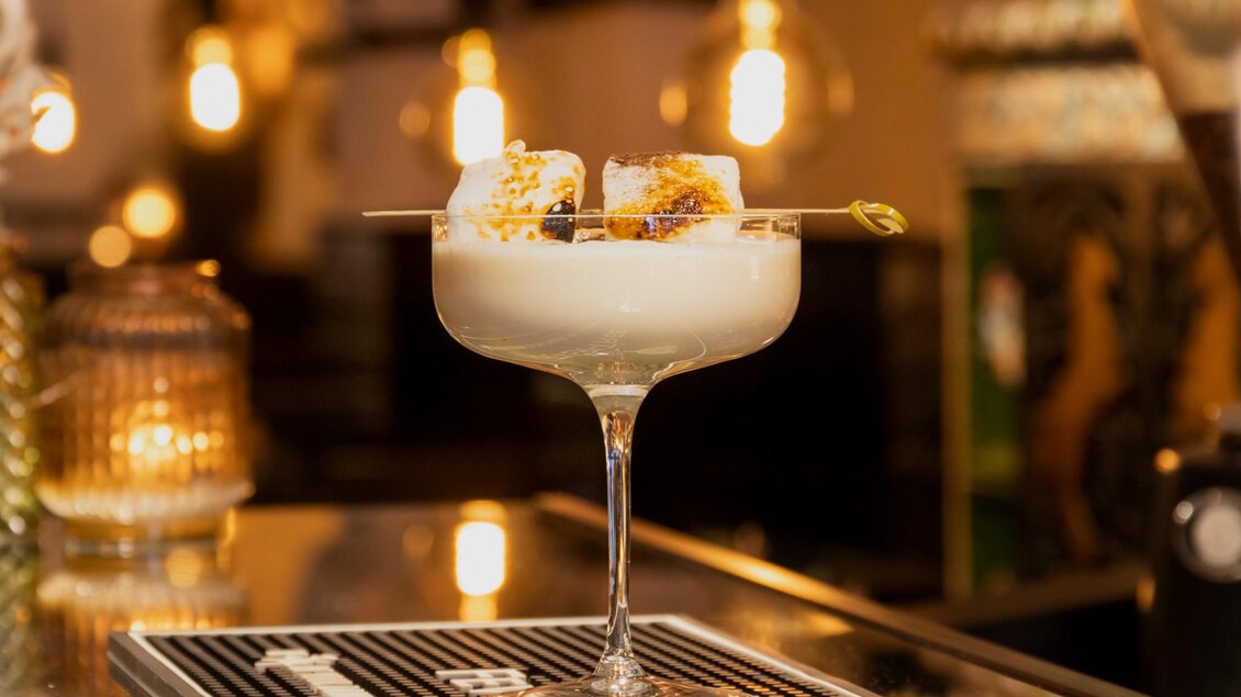 Ein eleganter Cocktail in einem hohen Glas, garniert mit Karamellbonbons. Im Hintergrund sind sanfte Lichter und eine stilvolle Bar zu sehen. | © Magdalena Bereznicka