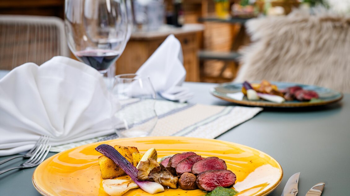 Ein elegantes Gericht auf einem gelben Teller mit Fleisch, Gemüse und einer Sauce. Im Hintergrund steht ein Weinglas und eine weitere Speise ist sichtbar. | © Region Graz, Mias Photoart