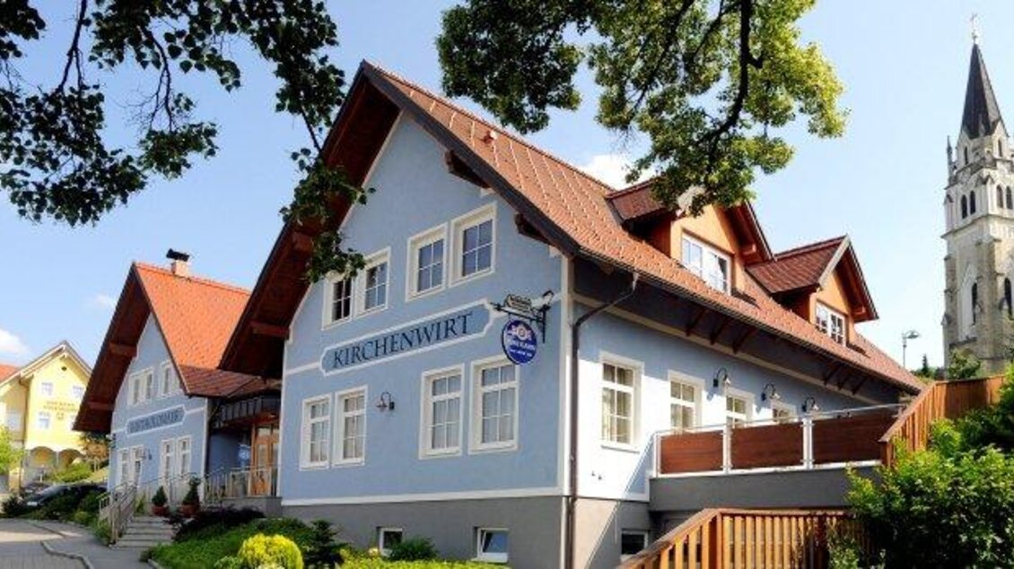 Ein charmantes Gasthaus mit blauem Anstrich und roten Dächern. Im Hintergrund ist ein Kirchturm sichtbar, umgeben von Bäumen. | © fb