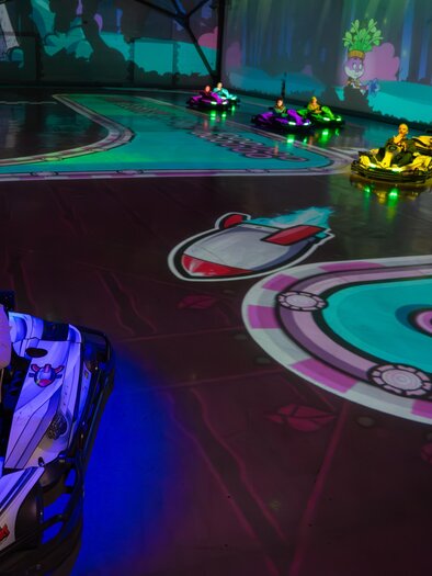 Drivers in electric karts on a hall with a brightly projected racetrack, colorful light plays, and digital game elements. | © Battlekart Gössendorf - Stefan Novotny