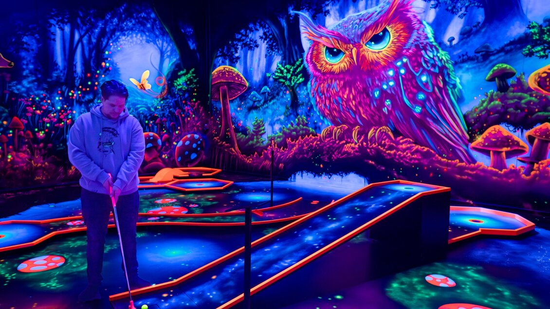 Person spielt Minigolf auf leuchtendem Blacklight-Parcours mit bunter Eulenwand, Neonfarben und fantasievoller Waldkulisse | © Battlekart Gössendorf - Stefan Novotny