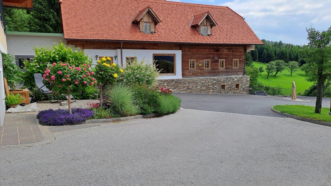 Ein schönes, rustikales Haus mit rotem Dach und grünen Wiesen im Hintergrund. Bunte Blumen schmücken den Weg zum Haus. | © Familienbetrieb Buchegger
