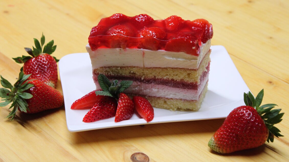 Ein Stück Erdbeertorte mit mehreren Schichten und frischen Erdbeeren. Die Torte hat eine glatte Creme und eine glänzende Erdbeerglasur. | © Bauernhofbackstube Rössing