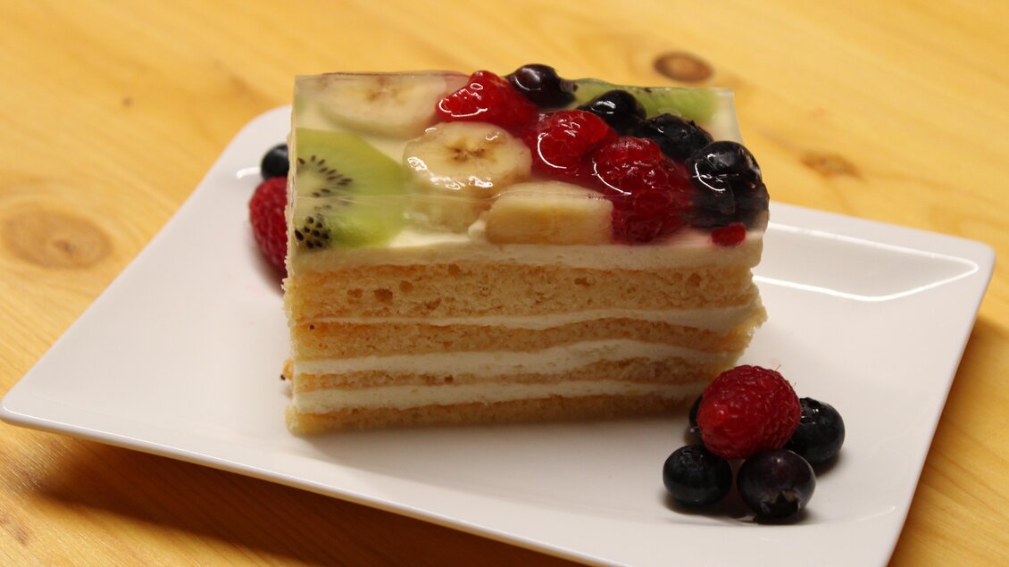 Eine köstliche Obsttorte mit mehreren Schichten und bunten Früchten. Serviert auf einem weißen Teller mit frischen Beeren. | © Bauernhofbackstube Rössing