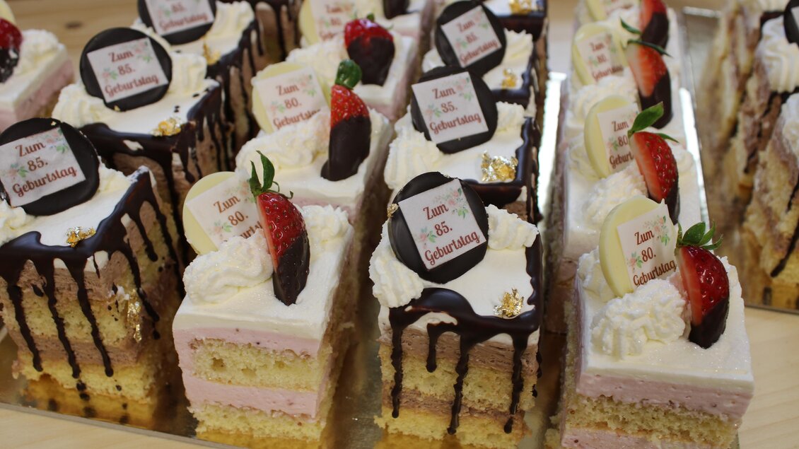 Eine köstliche Auswahl an Geburtstagskuchen mit Sahne, Schokolade und frischen Erdbeeren. Die Kuchensorten sind elegant angerichtet und mit Aufschriften versehen. | © Bauernhofbackstube Rössinger
