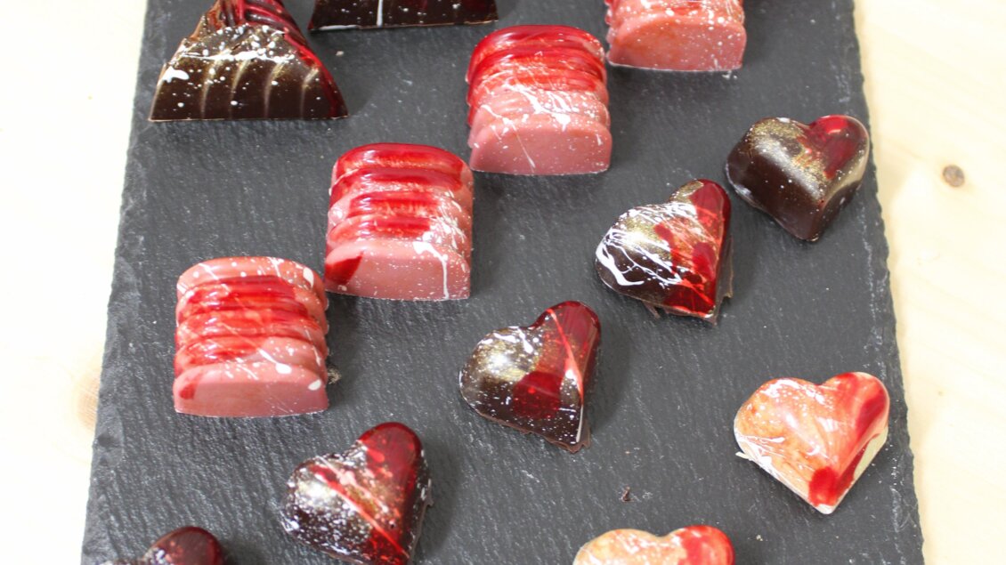 Ein schönes Arrangement von schokoladigen Pralinen in verschiedenen Formen, darunter Herzen und Pyramiden. Die Farben variieren von rot bis dunkelbraun auf einer eleganten schwarzen Platte. | © Bauernhofbackstube Rössinger