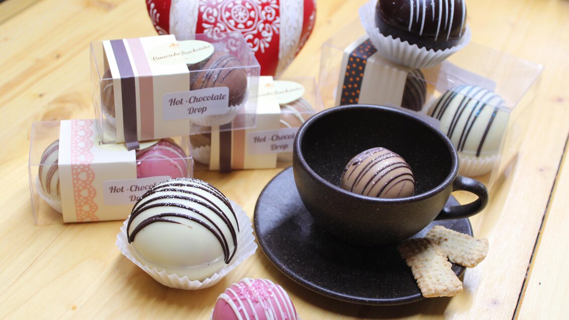 Ein stilvolles Arrangement mit verschiedenen Schokoladenkugeln und einer Tasse. Im Hintergrund ist ein rotes Herzdekor und einige Süßigkeiten zu sehen. | © Bauernhofbackstube Rössinger