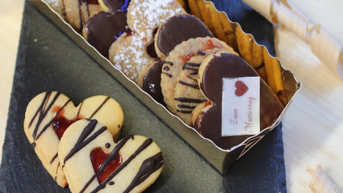 Eine ansprechende Auswahl von herzförmigen Keksen in verschiedenen Geschmacksrichtungen, liebevoll dekoriert. Im Hintergrund sind eine rote Rose und dekorative Elemente zu sehen. | © Bauernhofbackstube Rössinger