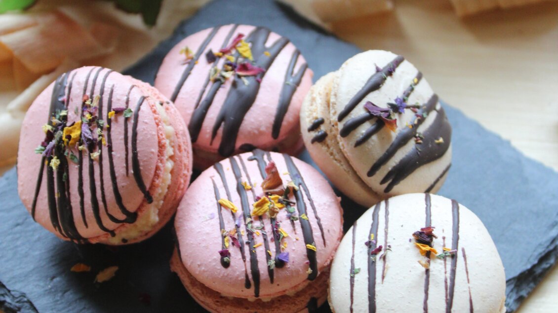 Eine Auswahl an bunten Macarons auf einem schwarzen Steinuntergrund. Die Süßigkeiten sind mit Schokoladensoße und Blumen dekoriert. | © Bauernhofbackstube Rössinger