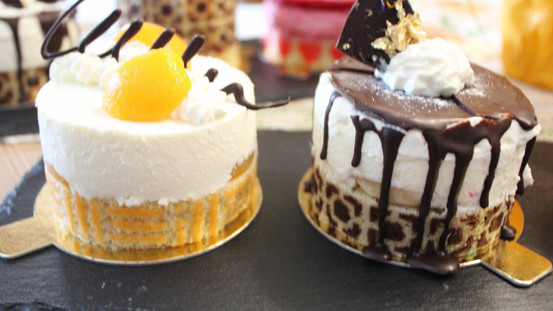 Zwei köstliche Kuchen auf einer schwarzen Platte. Der linke Kuchen hat eine Schicht aus Sahne und Pfirsich, während der rechte mit Schokolade und Sahne dekoriert ist. | © Bauernhofbackstube Rössinger