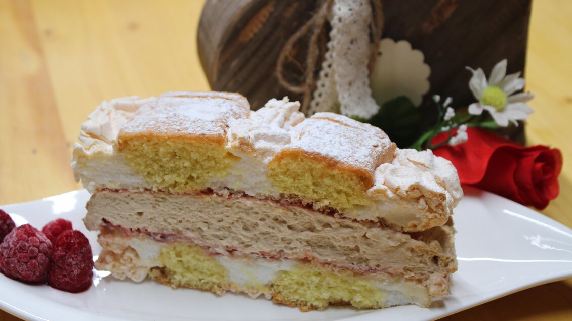 Ein köstlicher Kuchen mit mehreren Schichten, darunter Biskuit und Creme. Neben dem Kuchen liegen frische Himbeeren und eine rote Rose auf dem Teller. | © Bauernhofbackstube Rössinger