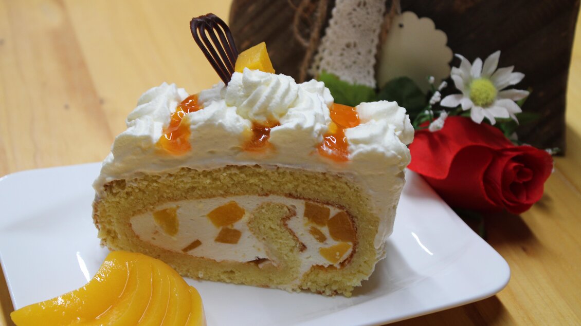 Ein Stück Biskuitrolle mit Sahne und Früchten, dekoriert mit einer Schokoladenspirale. Neben dem Kuchen liegen einige Pfirsichscheiben. | © Bauernhofbackstube Rössinger