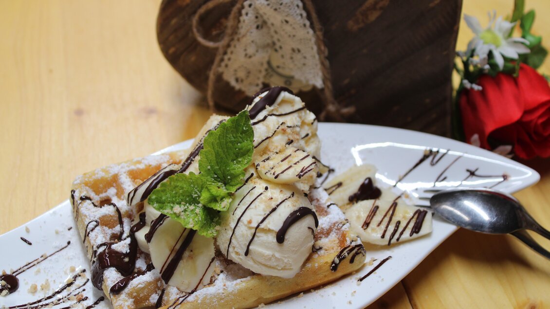 Ein köstliches Waffeldessert mit Vanilleeis, Bananen und Schokoladensauce. Es ist auf einem weißen Teller serviert und wird von einer Pflanzen-Dekoration begleitet. | © Bauernhofbackstube Rössinger