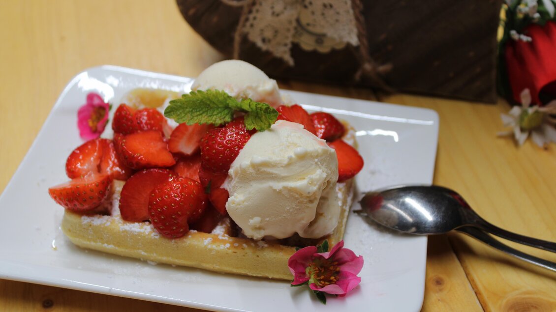 Ein Teller mit Waffeln, frischen Erdbeeren und zwei Kugeln Eis. Das Dessert ist mit Minzblättern und Blüten dekoriert. | © Bauernhofbackstube Rössinger