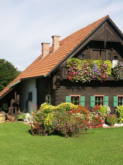 Ein charmantes, traditionelles Holzhaus mit bunten Blumen auf dem Balkon. Die grüne Wiese und die Bäume im Hintergrund schaffen eine einladende Atmosphäre.