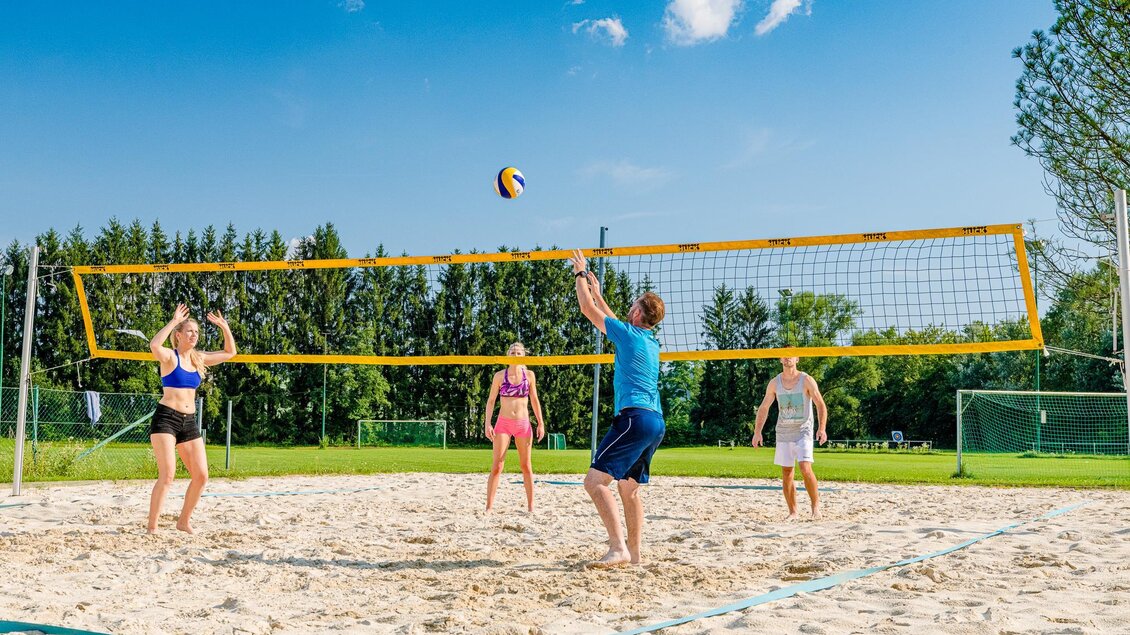Eine Gruppe junger Menschen spielt Beachvolleyball auf einem Sandplatz. Der Himmel ist blau mit einigen Wolken und die Umgebung ist grün. | © Mias Photoart